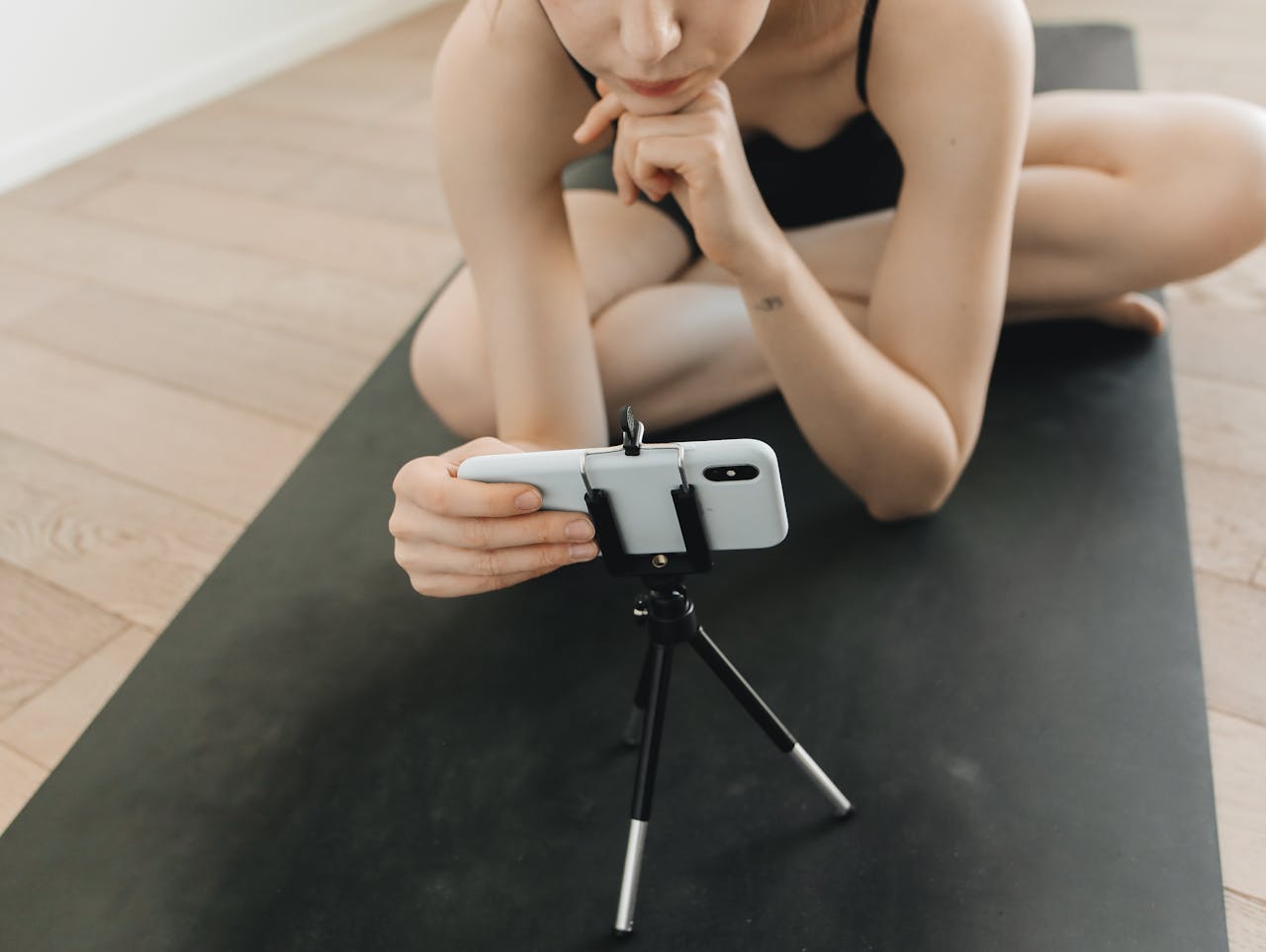 gallery-02 A woman in activewear vlogs using a smartphone and tripod while seated on a yoga mat indoors.